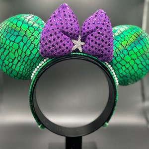 Op de afbeelding: Een paar muizenoren met groene, geschubde oren en een paarse paillettenstrik. De zwarte hoofdband is versierd met witte, parelachtige kralen. Een zilveren stervormige versiering bevindt zich in het midden van de strik.