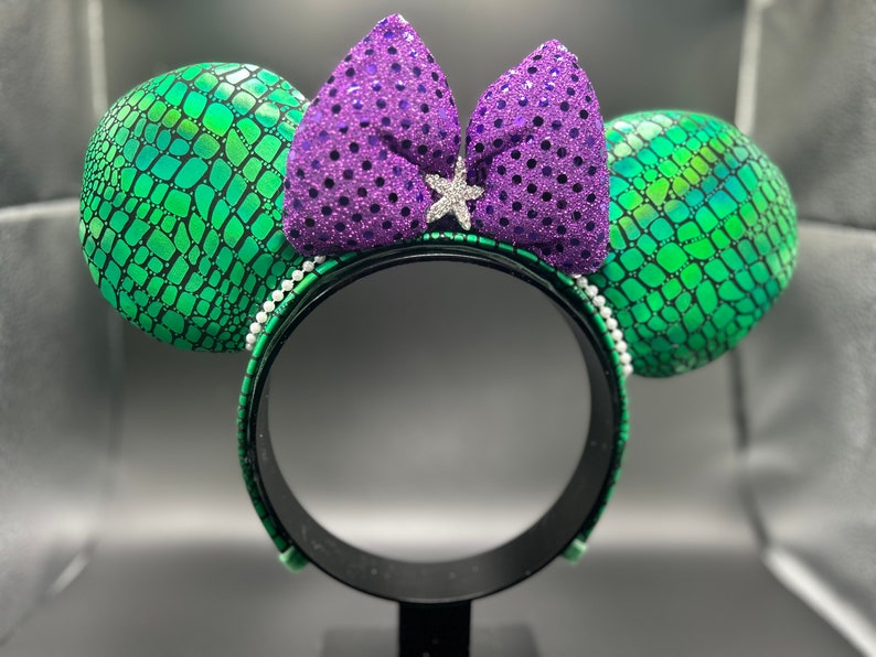 Op de afbeelding: Een paar muizenoren met groene, geschubde oren en een paarse paillettenstrik. De zwarte hoofdband is versierd met witte, parelachtige kralen. Een zilveren stervormige versiering bevindt zich in het midden van de strik.