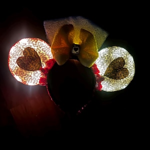 May include: A headband featuring two circular ears covered in shimmering gold sequins, each with a dark heart shape in the center. A large yellow bow sits between the ears, accented with a small square charm displaying a black heart symbol.