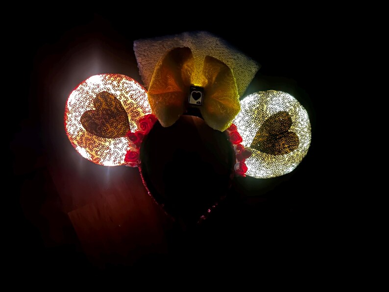 May include: A headband featuring two circular ears covered in shimmering gold sequins, each with a dark heart shape in the center. A large yellow bow sits between the ears, accented with a small square charm displaying a black heart symbol.