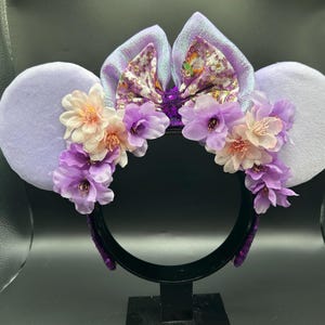 Puede incluir: Una diadema con dos orejas grandes y redondas de color violeta claro y un lazo central de tela iridiscente con estampado floral. La base de las orejas est&aacute; decorada con un arreglo de flores artificiales en tonos violeta y crema.