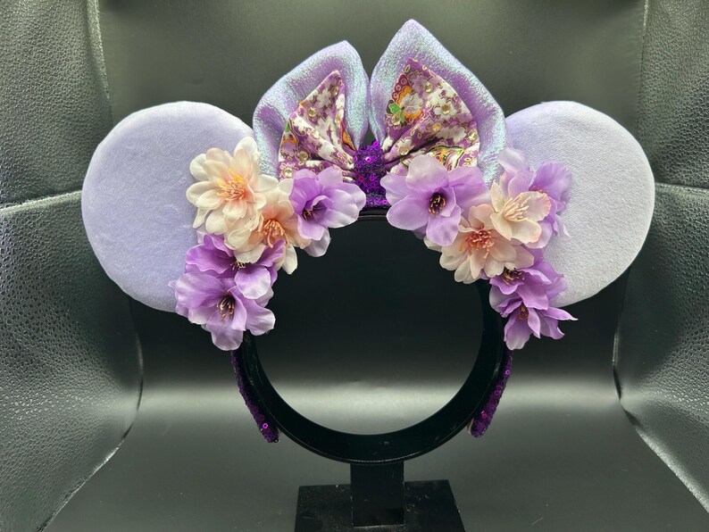Puede incluir: Una diadema con dos orejas grandes y redondas de color violeta claro y un lazo central de tela iridiscente con estampado floral. La base de las orejas est&aacute; decorada con un arreglo de flores artificiales en tonos violeta y crema.