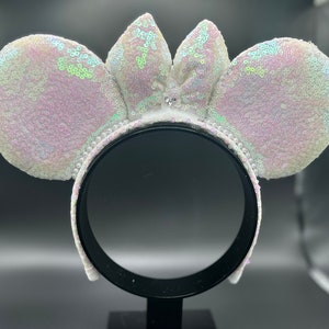 Puede incluir: Diadema de orejas de Minnie Mouse de lentejuelas blancas iridiscentes con un lazo blanco en el centro.