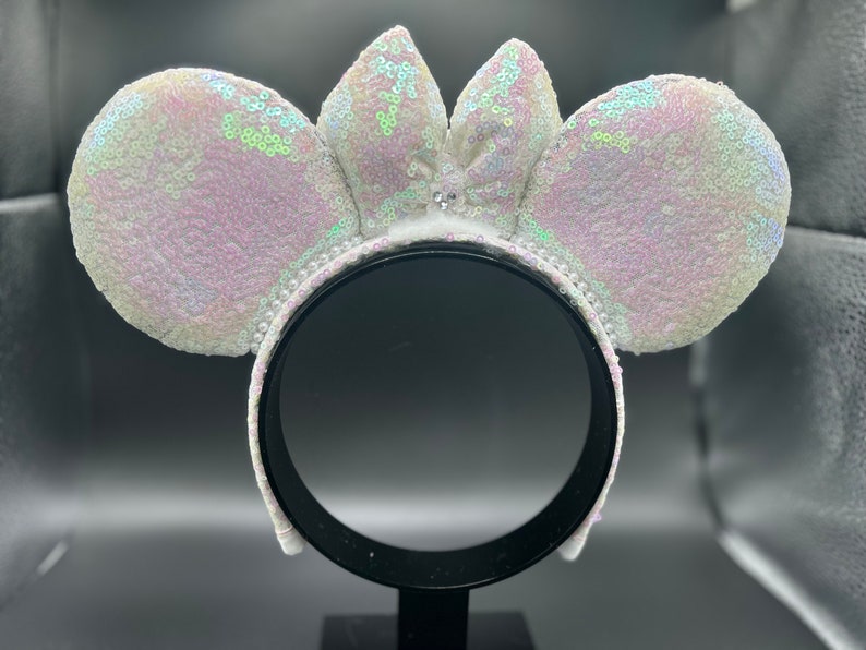 Puede incluir: Diadema de orejas de Minnie Mouse de lentejuelas blancas iridiscentes con un lazo blanco en el centro.