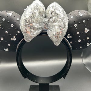 May include: Black headband with two round black ears adorned with clear rhinestones and small Mickey Mouse-shaped gems. A silver sequined bow sits between the ears. The headband is black.