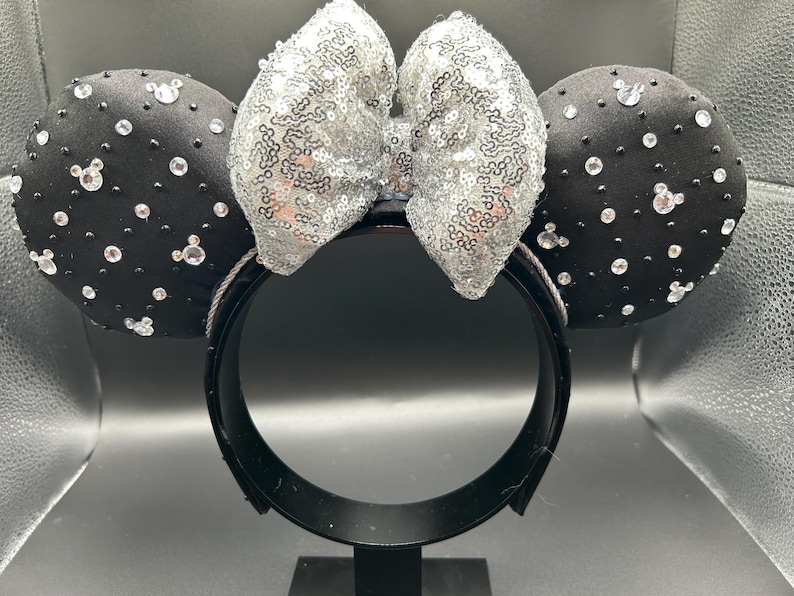 May include: Black headband with two round black ears adorned with clear rhinestones and small Mickey Mouse-shaped gems. A silver sequined bow sits between the ears. The headband is black.