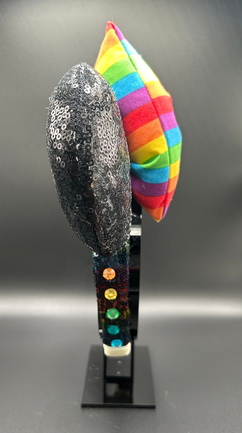 K&ouml;nnte beinhalten: Schwarze Pailletten-Minnie-Maus-Ohren mit einer regenbogenfarbenen Schleife aus Stoff. Das Stirnband ist mit regenbogenfarbenen Edelsteinen verziert.