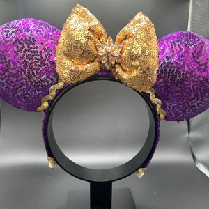 Pu&ograve; includere: Orecchie di Minnie Mouse con paillettes viola e dorate, con un grande fiocco dorato e un cristallo trasparente al centro.