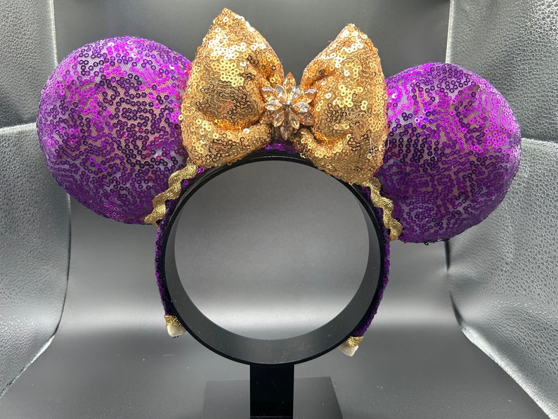 Pu&ograve; includere: Orecchie di Minnie Mouse con paillettes viola e dorate, con un grande fiocco dorato e un cristallo trasparente al centro.
