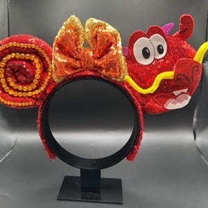 May include: Red sequined headband with a bow and dragon design. The headband features a large bow with orange and gold sequins. One ear is a spiral of red sequins with gold beads. The other ear is a red dragon with white eyes and a yellow mustache.