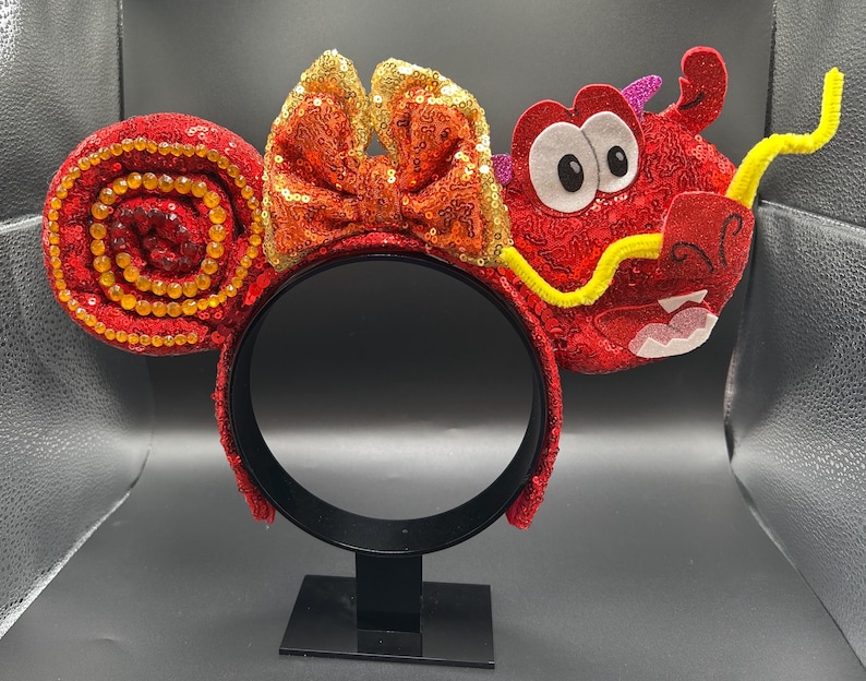 May include: Red sequined headband with a bow and dragon design. The headband features a large bow with orange and gold sequins. One ear is a spiral of red sequins with gold beads. The other ear is a red dragon with white eyes and a yellow mustache.