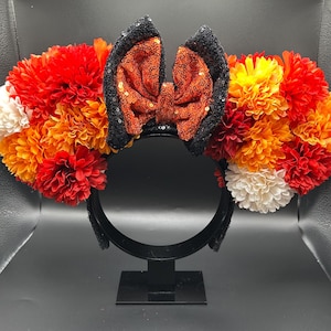 May include: A headband featuring two circular clusters of artificial flowers in shades of red, orange, yellow, and white. Centered between the floral clusters is a bow covered in orange sequins, flanked by two black sequined ear-shaped accents.