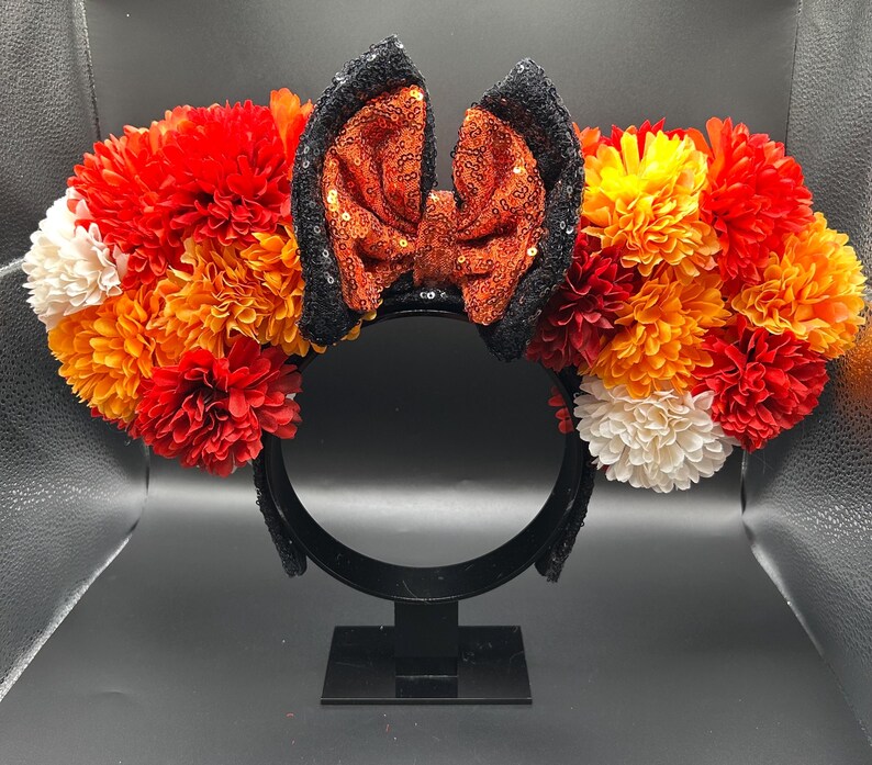 May include: A headband featuring two circular clusters of artificial flowers in shades of red, orange, yellow, and white. Centered between the floral clusters is a bow covered in orange sequins, flanked by two black sequined ear-shaped accents.