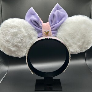 May include: A headband with large, fluffy white ears. A light purple velvet bow sits atop the headband, with a pink center adorned with a small jewel. The headband is white with silver trim, and the ears are soft and plush.