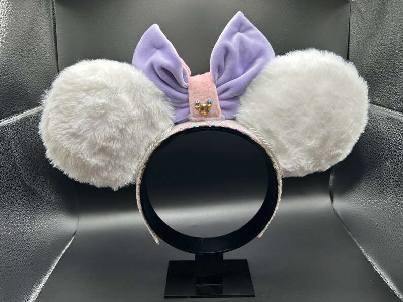 May include: A headband with large, fluffy white ears. A light purple velvet bow sits atop the headband, with a pink center adorned with a small jewel. The headband is white with silver trim, and the ears are soft and plush.