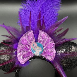 May include: A headband featuring two black sequined ears, a central lavender sequined bow, and a vibrant purple feather plume. A circular enamel pin depicting a stylized character with light skin and flowing hair is centered on the bow.