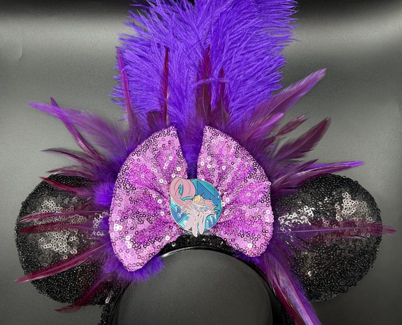 May include: A headband featuring two black sequined ears, a central lavender sequined bow, and a vibrant purple feather plume. A circular enamel pin depicting a stylized character with light skin and flowing hair is centered on the bow.