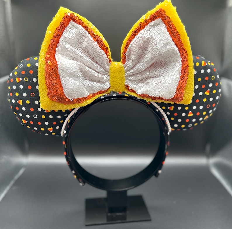 May include: A black headband with two round ears and a large bow. The ears are black with white, orange, and yellow polka dots. The bow is white with sequins, outlined in orange and yellow.