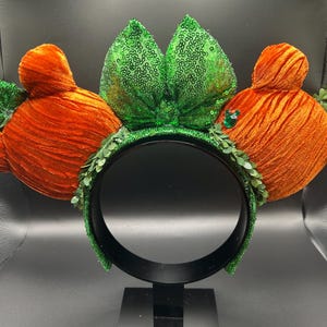 May include: A whimsical headband featuring orange velvet carrot-shaped ears with green sequined leaves and a green sequined bow. The headband is adorned with green leaf accents and a black band.