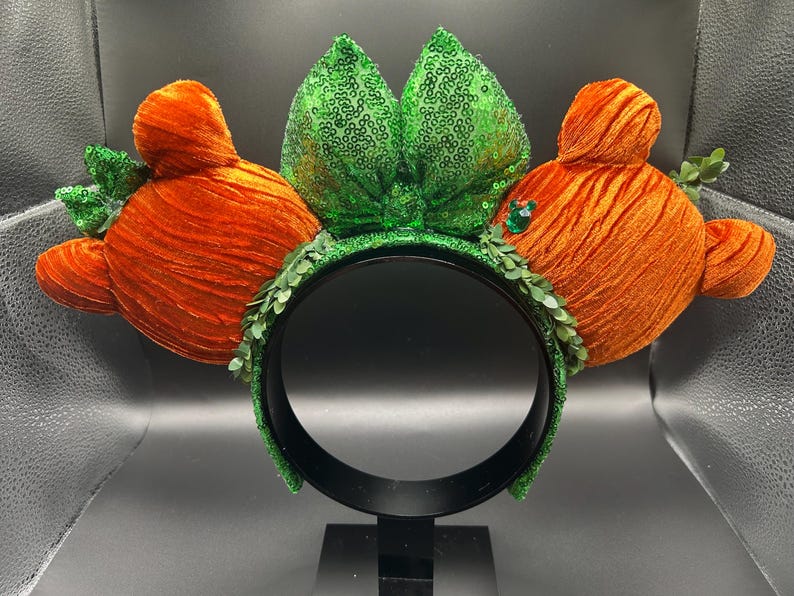May include: A whimsical headband featuring orange velvet carrot-shaped ears with green sequined leaves and a green sequined bow. The headband is adorned with green leaf accents and a black band.