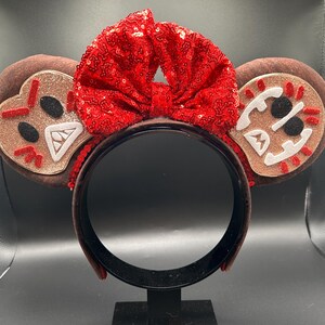 May include: A headband featuring two round brown ears with glittery tan centers. Each ear displays a stitched face with black eyes, white teeth, and red accents. A large, bright red sequined bow is centered between the ears on the headband.