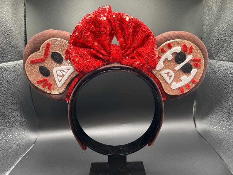 May include: A headband featuring two round brown ears with glittery tan centers. Each ear displays a stitched face with black eyes, white teeth, and red accents. A large, bright red sequined bow is centered between the ears on the headband.