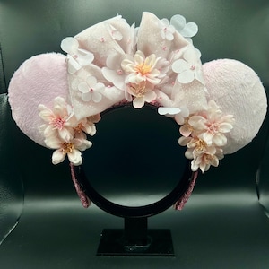 Op de afbeelding: Een haarband met twee ronde, lichtroze, pluizige oren en een grote, glinsterende, lichtroze stoffen strik in het midden. Het ontwerp is versierd met talloze kleine, delicate witte en lichtroze bloemaccenten.