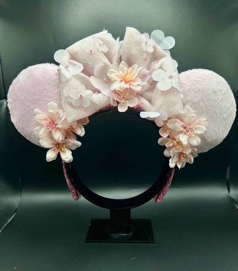 Op de afbeelding: Een haarband met twee ronde, lichtroze, pluizige oren en een grote, glinsterende, lichtroze stoffen strik in het midden. Het ontwerp is versierd met talloze kleine, delicate witte en lichtroze bloemaccenten.