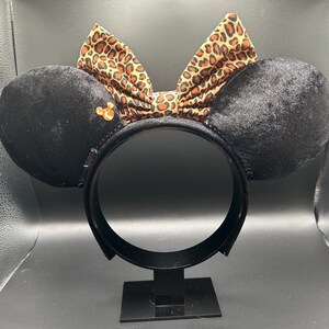 以下が含まれることがあります： 黒いベルベットのミッキーマウスの耳に、ヒョウ柄のリボンと小さな金色の装飾が施されています。耳は黒いヘッドバンドに取り付けられています。リボンは茶色とタンのアニマルプリント柄です。耳は黒いスタンドに展示されています。
