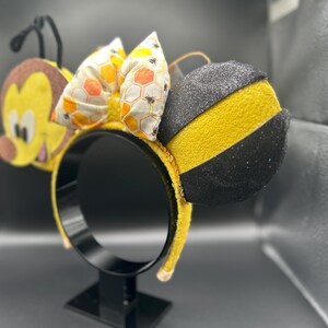 May include: Yellow and black bee-themed Minnie Mouse ears with a honeycomb bow. The ears are made of felt and have a black glitter finish.