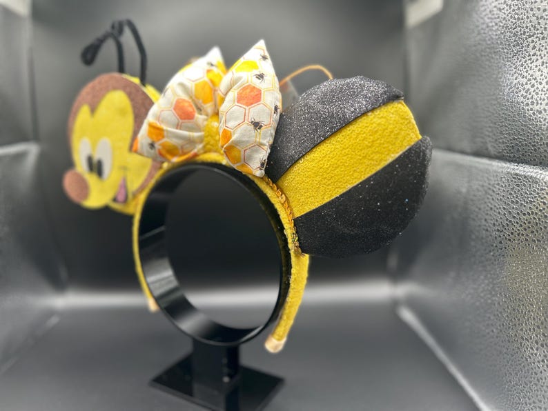 May include: Yellow and black bee-themed Minnie Mouse ears with a honeycomb bow. The ears are made of felt and have a black glitter finish.