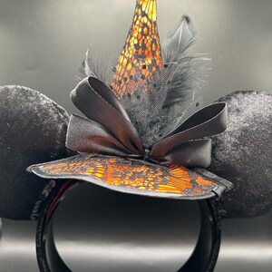 Puede incluir: Una diadema con dos grandes orejas redondas de terciopelo negro y un sombrero de bruja central. El sombrero es naranja con una capa de encaje negro, acentuado por un lazo de sat&eacute;n negro, tul negro y plumas negras en la base.