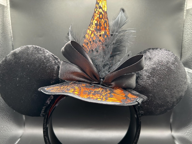 Puede incluir: Una diadema con dos grandes orejas redondas de terciopelo negro y un sombrero de bruja central. El sombrero es naranja con una capa de encaje negro, acentuado por un lazo de sat&eacute;n negro, tul negro y plumas negras en la base.