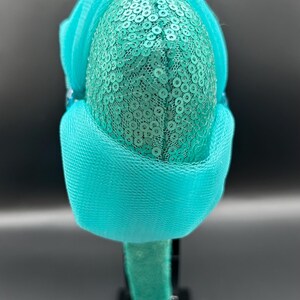 May include: A teal sequined headband with a gold accent. The headband features a teal mesh overlay and is set against a dark background. The design is centered on a black stand.