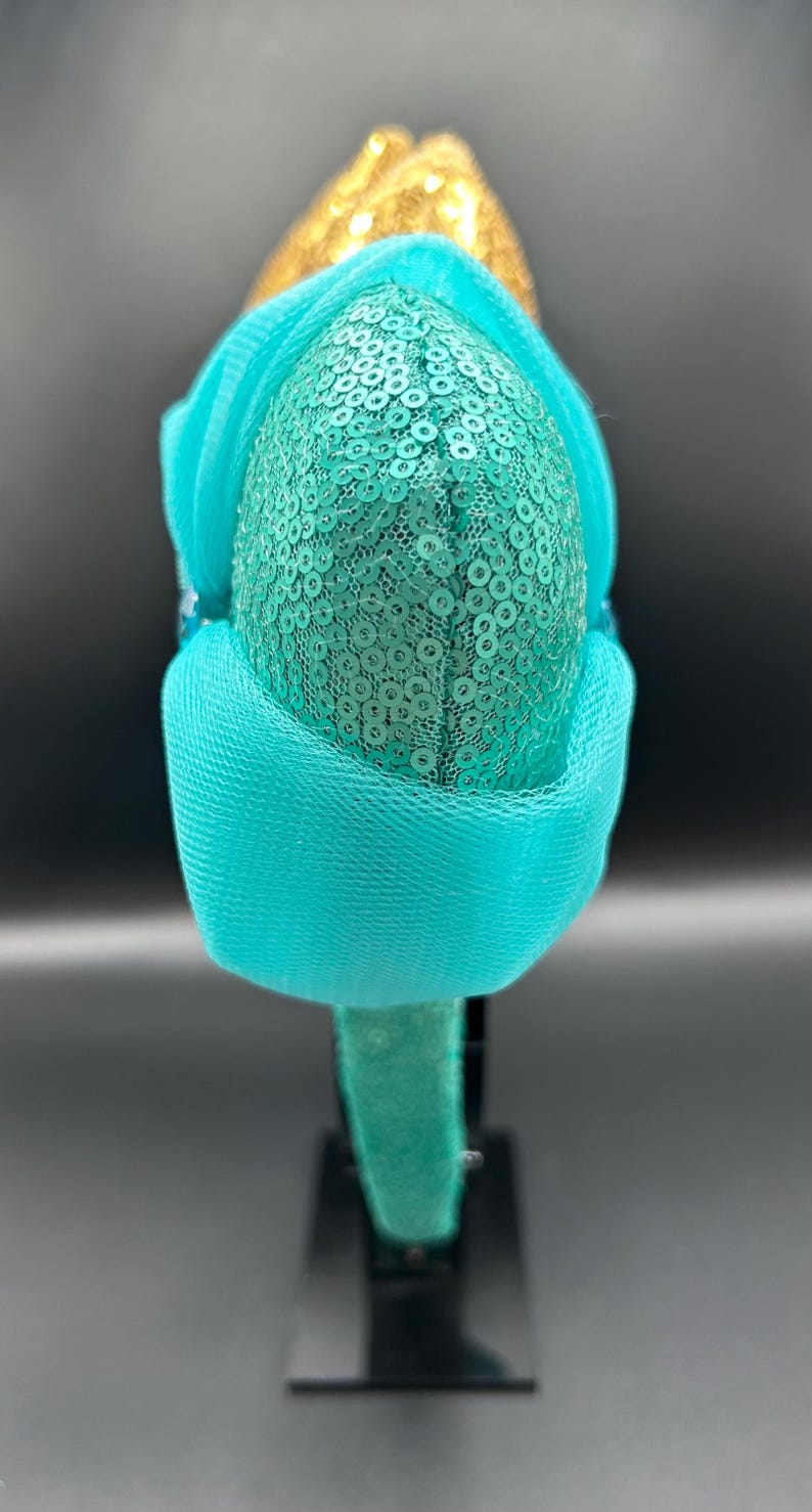 May include: A teal sequined headband with a gold accent. The headband features a teal mesh overlay and is set against a dark background. The design is centered on a black stand.