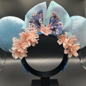 Peut inclure: Un serre-t&ecirc;te avec deux grandes oreilles en velours bleu clair, un n&oelig;ud central bleu clair iris&eacute; et un n&oelig;ud en tissu violet &agrave; motif floral. Le serre-t&ecirc;te est orn&eacute; de grappes de fleurs de cerisier artificielles rose p&acirc;le.