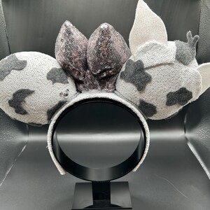 May include: A headband featuring two round ears covered in light grey textured fabric with dark grey patches. A central bow is adorned with shimmering dark grey sequins. The right ear includes two small, light grey pointed fabric accents.