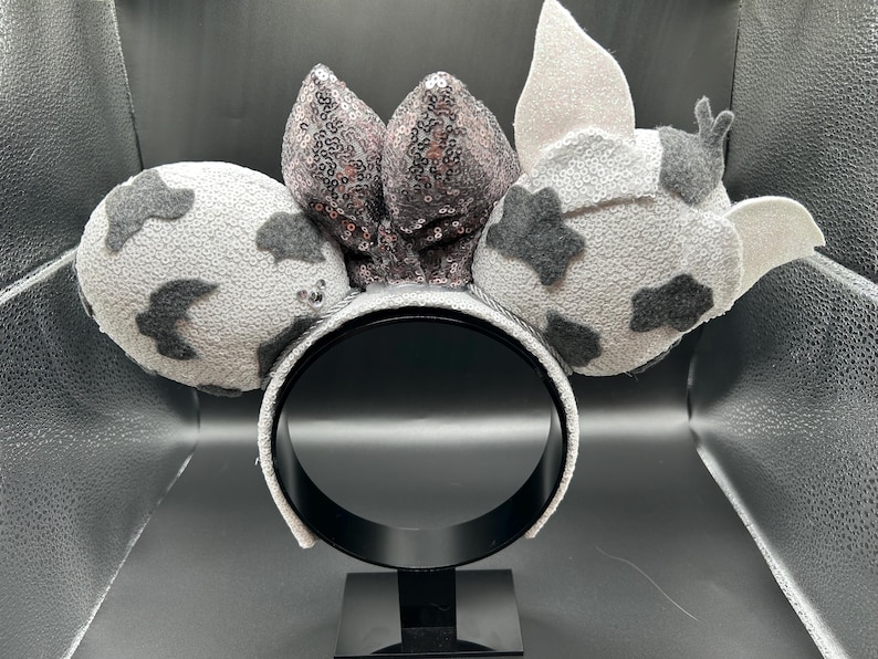 May include: A headband featuring two round ears covered in light grey textured fabric with dark grey patches. A central bow is adorned with shimmering dark grey sequins. The right ear includes two small, light grey pointed fabric accents.