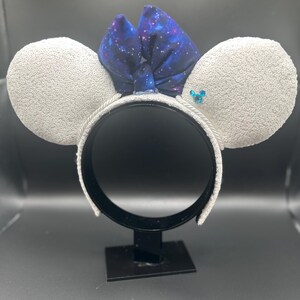 May include: White sequined mouse ear headband with a galaxy print bow. The ears are covered in white sequins, and the bow features a dark blue fabric with white stars. A small blue gem is on one ear.