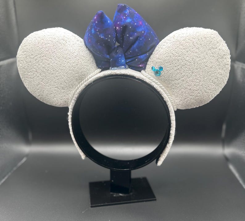 May include: White sequined mouse ear headband with a galaxy print bow. The ears are covered in white sequins, and the bow features a dark blue fabric with white stars. A small blue gem is on one ear.