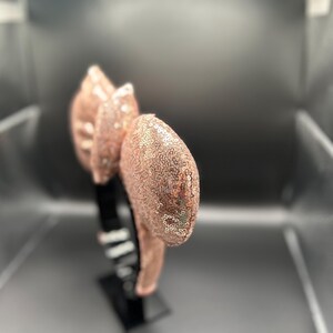 May include: A rose gold sequined headband with large, rounded ears. The ears and bow are covered in shimmering sequins. The headband is black and appears to be made of plastic. The accessory is displayed against a gray background.