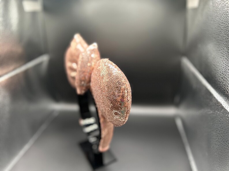 May include: A rose gold sequined headband with large, rounded ears. The ears and bow are covered in shimmering sequins. The headband is black and appears to be made of plastic. The accessory is displayed against a gray background.