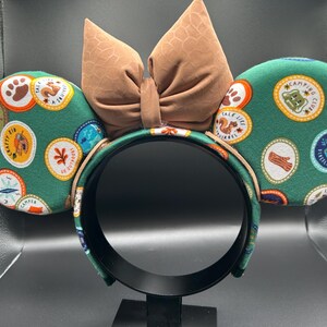 May include: Green and brown mouse ears with a brown bow. The ears are covered in a pattern of colourful patches with text that says "Camp", "Talk Like a Squirrel", "Shine Brightly", and more.