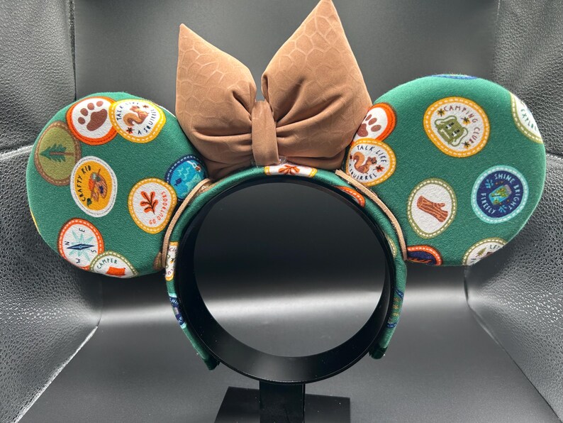 May include: Green and brown mouse ears with a brown bow. The ears are covered in a pattern of colourful patches with text that says "Camp", "Talk Like a Squirrel", "Shine Brightly", and more.