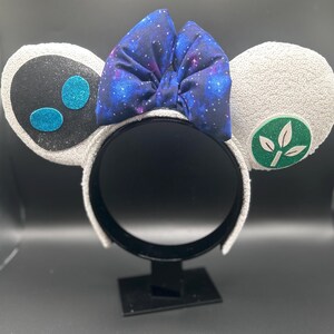 May include: White sequined mouse ear headband with a galaxy print bow. One ear features a black face with blue glitter accents, the other a green leaf design. A fun accessory for themed events.