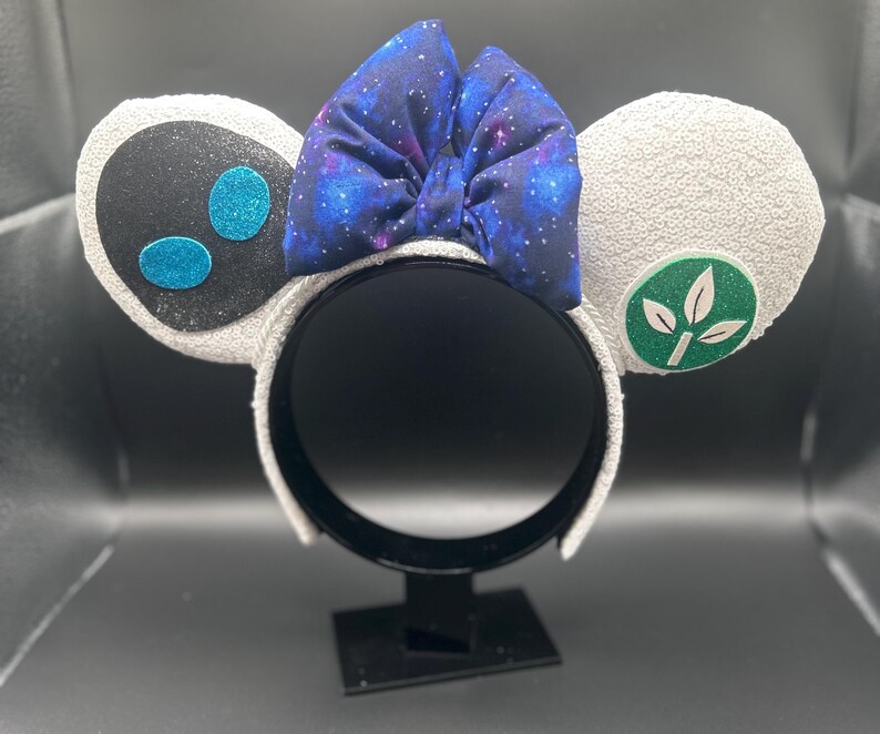 May include: White sequined mouse ear headband with a galaxy print bow. One ear features a black face with blue glitter accents, the other a green leaf design. A fun accessory for themed events.