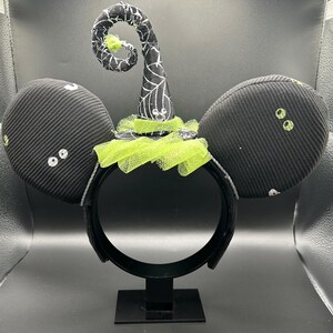 May include: Black Halloween-themed mouse ear headband with a witch hat. The ears are black with white accents and small, green, sequined eyes. The witch hat is black with white spiderweb detailing and a green tulle trim. The headband is black.