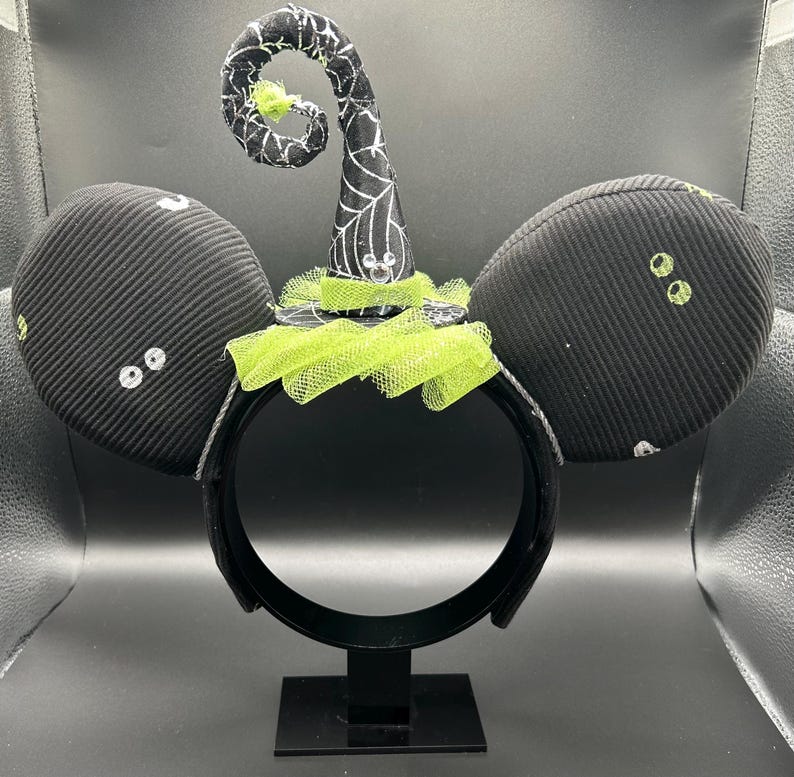 May include: Black Halloween-themed mouse ear headband with a witch hat. The ears are black with white accents and small, green, sequined eyes. The witch hat is black with white spiderweb detailing and a green tulle trim. The headband is black.