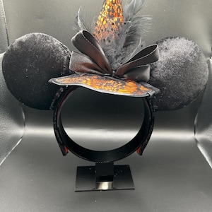 Puede incluir: Una diadema con dos grandes orejas circulares de terciopelo negro y un sombrero de bruja central. El sombrero es naranja con una capa de encaje negro, decorado con un lazo de sat&eacute;n negro y plumas negras. La estructura es de color negro.