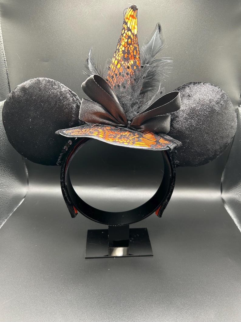 Puede incluir: Una diadema con dos grandes orejas circulares de terciopelo negro y un sombrero de bruja central. El sombrero es naranja con una capa de encaje negro, decorado con un lazo de sat&eacute;n negro y plumas negras. La estructura es de color negro.